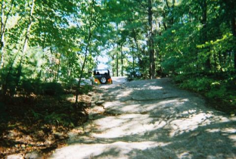 1976 GMC C/K Pickup - An off-road vehicle navigates a challenging rocky path through a wooded area.