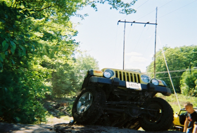 Yellow Jeep Wrangler tilted on a dirt path, front grille and headlights visible. License plate reads 94986