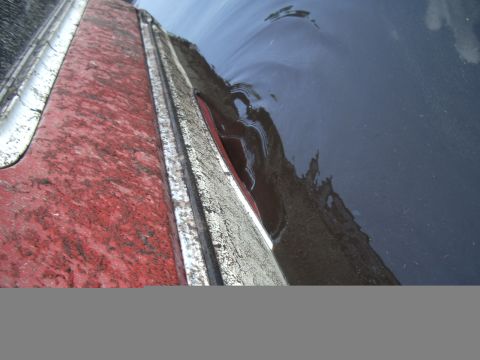 1978 Ford Bronco - A close-up view of a vehicle's body panel, showing a red section and a silver trim piece, with water reflecting on a dark surface.