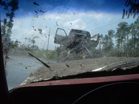1978 Ford Bronco - A monster truck is stuck in the mud, viewed from inside another vehicle with a dirty windshield.
