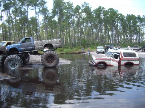1978 Ford Bronco - A red and white 1978 Ford Bronco is partially submerged in water, with a lifted pickup truck nearby.