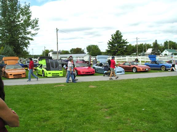 Orange, lime green, red, and blue 1984 Pontiac Fieros parked on grass with people walking by