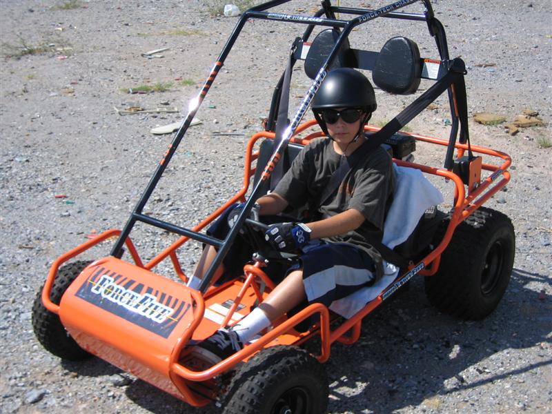 Kid wearing a helmet and sunglasses drives an orange go-kart with 'Force Five' on the front