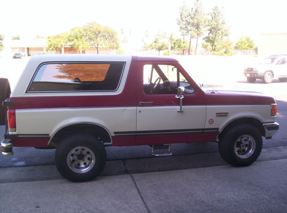 Side view of a red and white 1989 Ford Bronco with a spare tire on the back