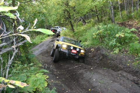 2003 Jeep TJ - A yellow Jeep Wrangler TJ navigates a challenging muddy trail in a wooded area, with other vehicles following.