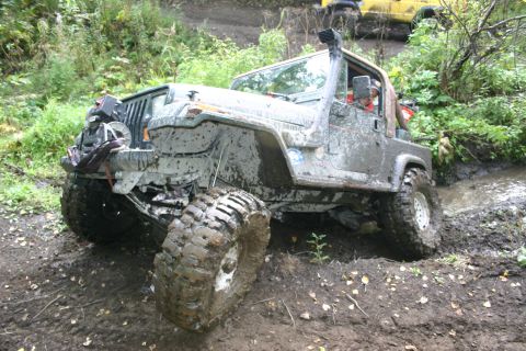 2003 Jeep TJ - A heavily muddied Jeep Wrangler drives through a water obstacle on an off-road trail, with another vehicle visible in the background.