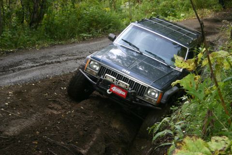 2003 Jeep TJ - A dark blue Jeep Cherokee XJ, modified for off-road use, tackles a challenging muddy trail with one wheel significantly elevated.
