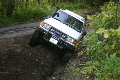 2003 Jeep TJ - White off-road SUV with snorkel and winch navigating a muddy ditch on a dirt road.