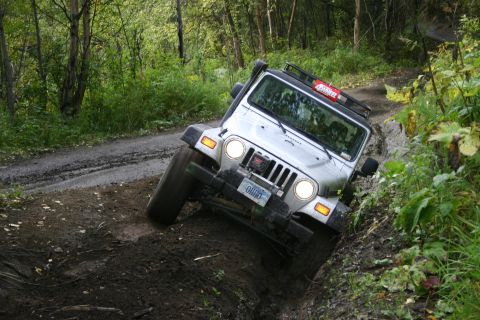 2003 Jeep TJ - A Jeep TJ navigates a challenging, muddy off-road path surrounded by dense foliage.