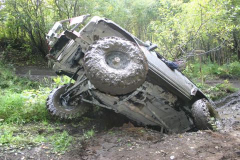 2003 Jeep TJ - An off-road vehicle is stuck in the mud, tipped onto its side on a trail surrounded by trees.