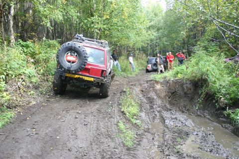 2003 Jeep TJ - Several off-road vehicles navigate a muddy trail through a forest, with one SUV appearing to be stuck.