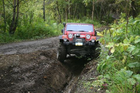 2003 Jeep TJ - A red 2003 Jeep TJ navigates a challenging muddy path in a forest setting.