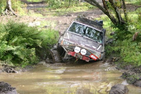 2003 Jeep TJ - A maroon Jeep Cherokee XJ navigates a muddy obstacle course, its front tires deep in the water.