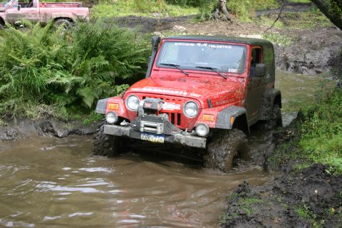 2003 Jeep TJ - This red Jeep Wrangler Rubicon navigates a challenging muddy trail, with another vehicle partially visible in the background.