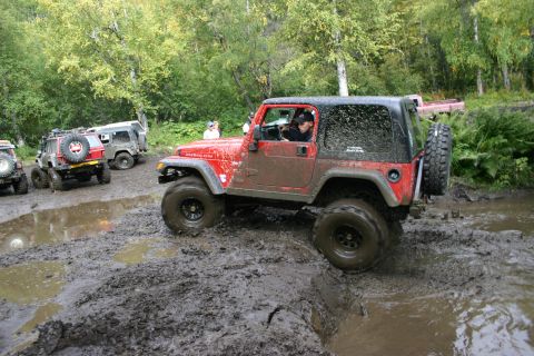 2003 Jeep TJ - A red 2003 Jeep TJ navigates a muddy off-road course, with other off-road vehicles visible in the background.
