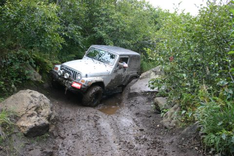 2003 Jeep TJ - A silver Jeep TJ tackles a challenging muddy incline surrounded by lush green vegetation and large boulders.