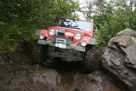 2003 Jeep TJ - A red 2003 Jeep TJ tackles a challenging muddy and rocky terrain, demonstrating its off-road capabilities.