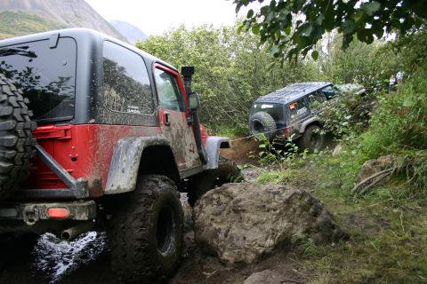 2003 Jeep TJ - Two Jeeps navigate a challenging muddy trail with large rocks and dense foliage.