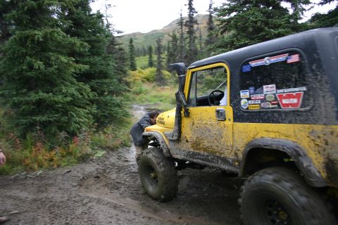 2003 Jeep TJ - A mud-covered yellow Jeep Wrangler is stopped on a dirt trail, with a person attending to the front of the vehicle.