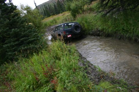 2003 Jeep TJ - A Jeep Wrangler navigates a water-filled, muddy trail surrounded by lush green vegetation.