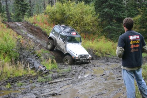 2003 Jeep TJ - A white Jeep Wrangler tackles a challenging muddy incline on an off-road trail, observed by a bystander.