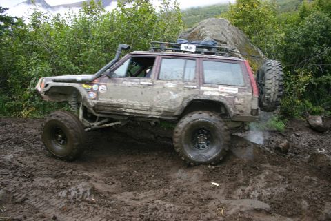 2003 Jeep TJ - A heavily modified Jeep Cherokee XJ navigates a muddy trail with a spare tire mounted on the back.
