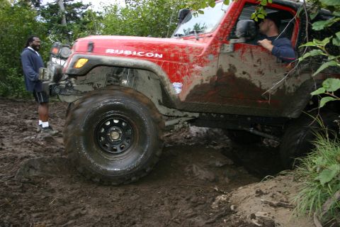 2003 Jeep TJ - A red 2003 Jeep TJ Rubicon navigates a muddy off-road trail with a driver and passenger.