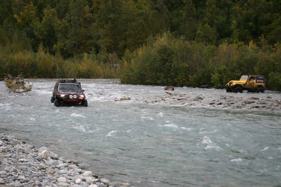 Brown Jeep TJ driving through a shallow river, front view with auxiliary lights. Yellow Jeep TJ visible in background