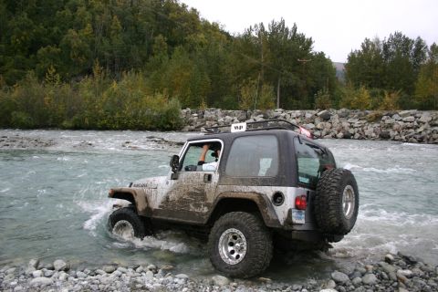 2003 Jeep TJ - A mud-splattered Jeep TJ navigates a rocky riverbed, its tires churning water.