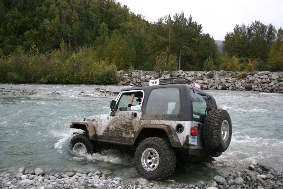 Silver Jeep TJ driving through a shallow river, covered in mud, with a black soft top and spare tire on the rear