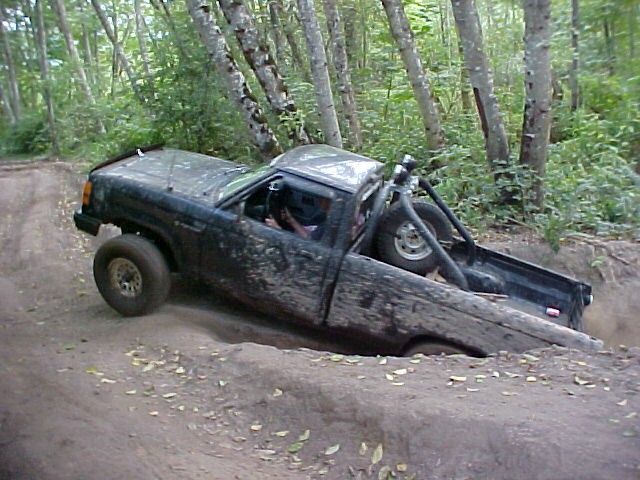 Muddy pickup truck stuck in a ditch on a wooded trail. It has a roll cage and a spare tire in the bed