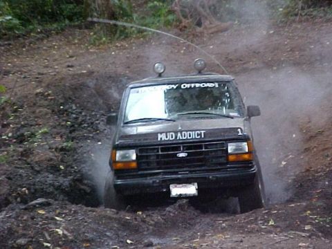 1992 Ford Ranger - A black SUV with "MUD ADDICT" and "HARDBOY OFFROAD" decals drives through a muddy off-road course.