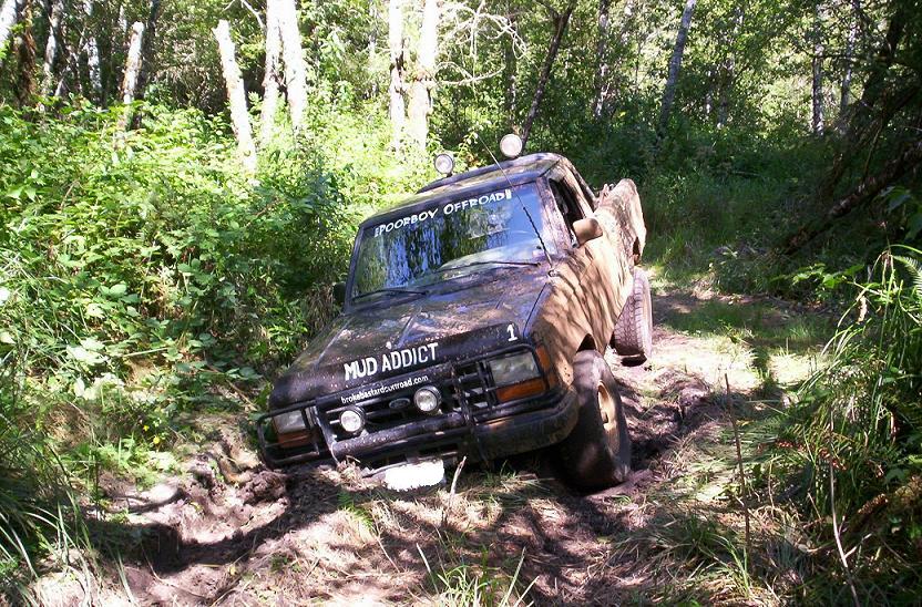Pickup truck stuck in mud on a wooded trail. It has 'MUD ADDICT' and 'POORBOY OFFROAD' stickers