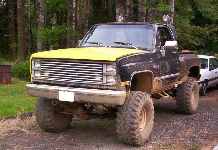Lifted black pickup truck with yellow hood, large off-road tires, and a white car parked behind it