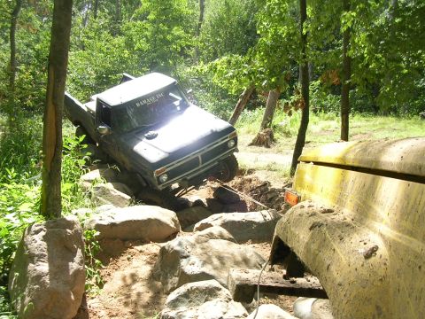 1977 Ford F-150 - A dark pickup truck navigates a challenging rocky path, with another yellow vehicle partially visible to the right.