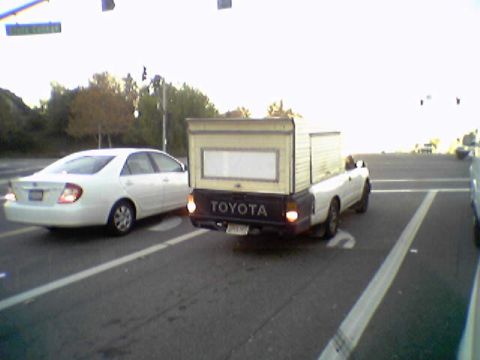 1990 Ford Bronco - Toyota pickup truck with a camper shell is stopped at a traffic light next to a white sedan.
