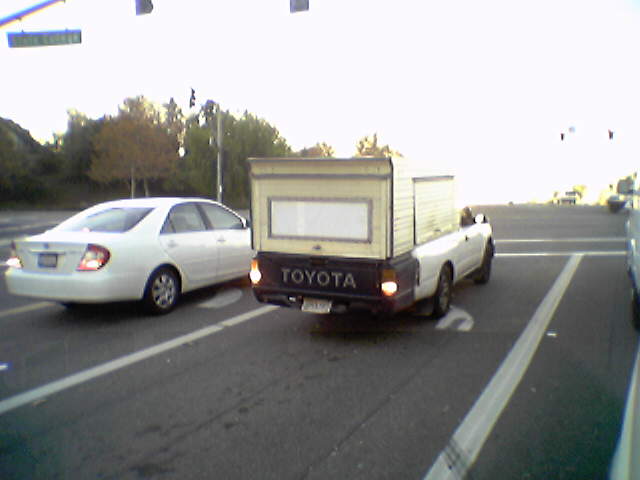 Toyota pickup truck with camper shell, white sedan in front. Traffic intersection