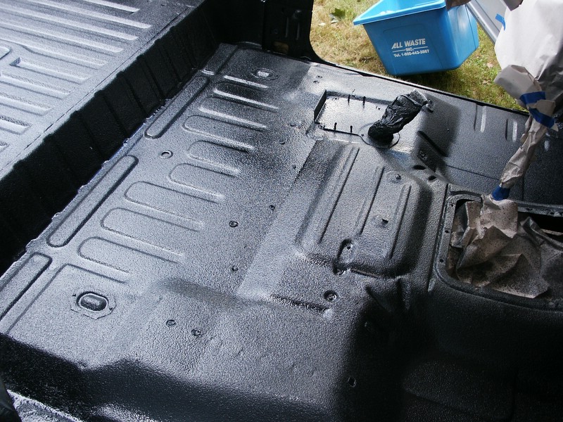 Vehicle interior floor with spray-in liner applied, showing ribbed texture and mounting points. Blue trash bin visible outside