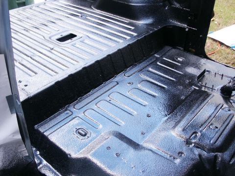 1982 Ford Bronco - The interior floor of a vehicle has been coated with a black spray-in liner, showing ribbed metal panels and a central transmission tunnel.