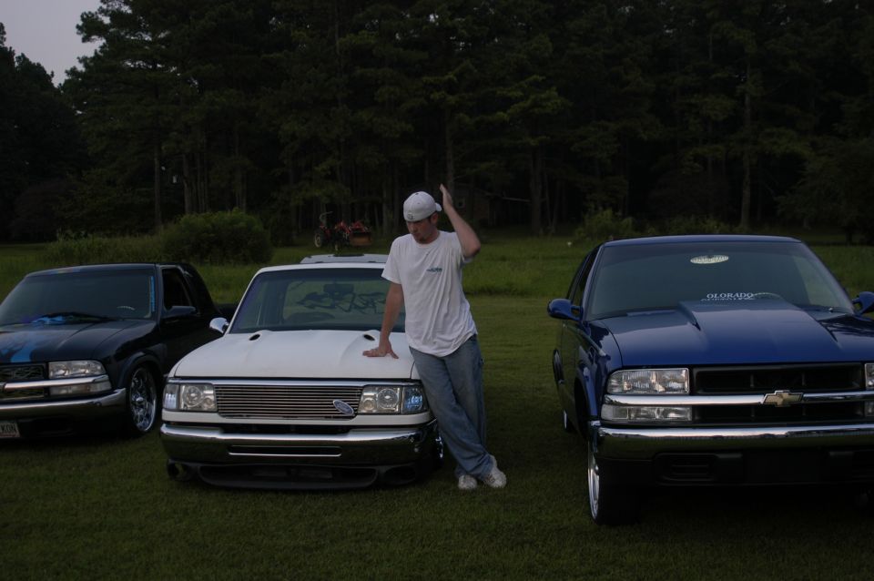 White pickup truck with a chrome grille and a man leaning on its hood, parked on grass with two other pickup trucks