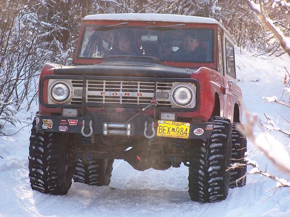 Red Ford Bronco with large tires driving in snow. License plate reads BEX 984