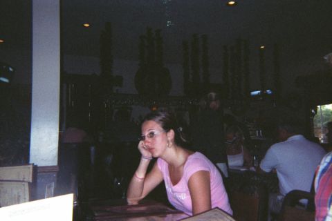 1998 Ford Taurus - A woman in a pink shirt and glasses sits at a table, resting her head on her hand, in a dimly lit indoor setting.