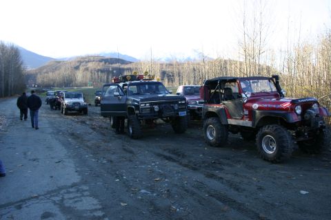 2003 Jeep TJ - A group of modified off-road vehicles are parked on a dirt road, with people walking nearby.
