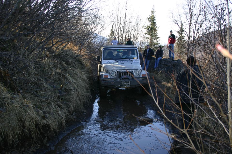 Silver 2003 Jeep TJ driving through a muddy, watery trail with people standing on a rock nearby
