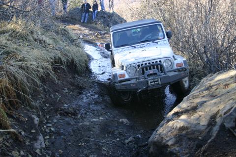 2003 Jeep TJ - A white 2003 Jeep TJ tackles a challenging muddy and rocky off-road course, with onlookers observing from a higher elevation.