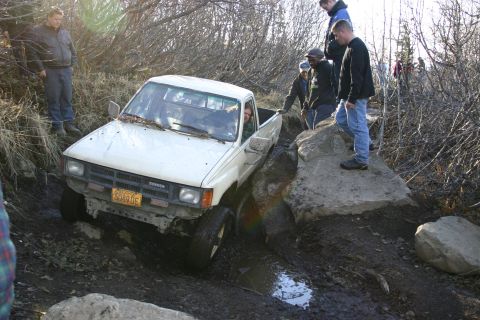 2003 Jeep TJ - White Toyota pickup truck attempts a difficult off-road obstacle with onlookers present.