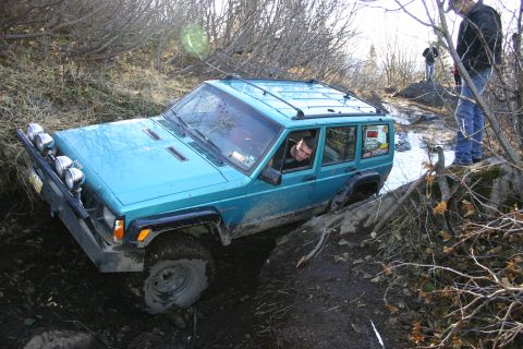 2003 Jeep TJ - A teal Jeep Cherokee XJ navigates a muddy trail with a driver visible through the window.
