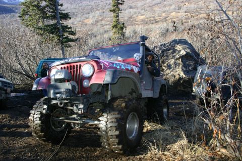 2003 Jeep TJ - A red Jeep Wrangler navigates a muddy, wooded trail with other vehicles nearby.