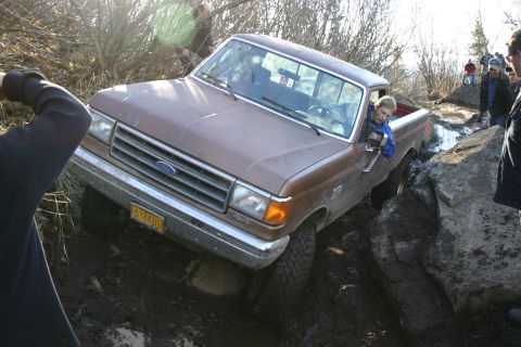 2003 Jeep TJ - A brown 1987 Ford F-150 pickup truck is shown off-roading through a rocky and muddy trail with several people observing.