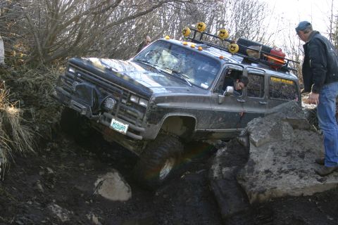 2003 Jeep TJ - A black SUV is shown off-roading on a muddy and rocky trail with people observing.